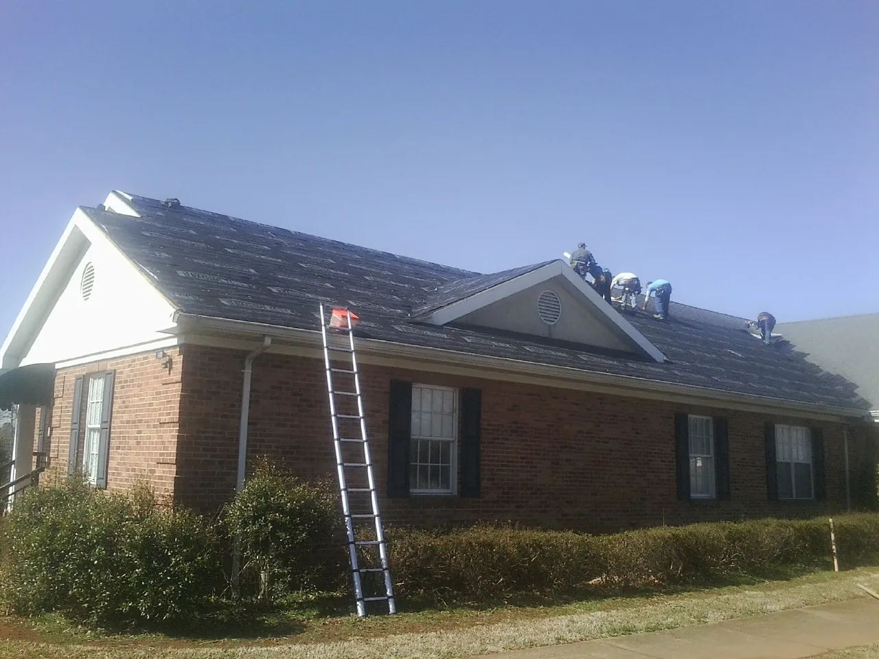 Crew tearing shingles off a brick ranch roof with a ladder set — Metal Roof Repair work by our crew in Angola